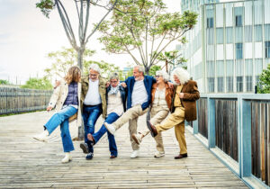 A group of elderly adults with their arms wrapped around each other's shoulders, kicking up their legs in a dance.