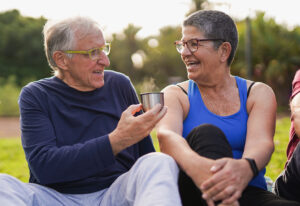 A man and a woman are sitting outside in the nice weather, and the man is offering the woman a steaming cup liquid.