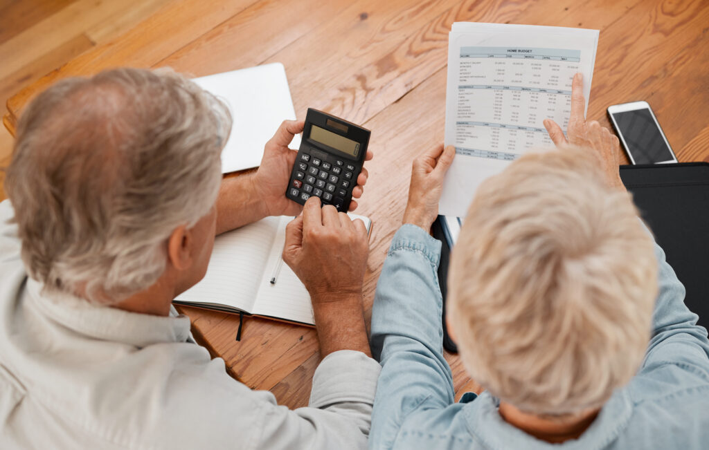 Retired couple calculating monthly home budget with bills and calculator at wooden table