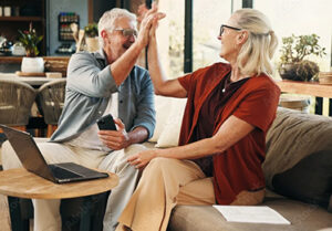 Mature couple sitting together at a table with a laptop and paperwork, smiling and celebrating with a high five.
