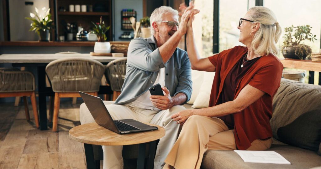 Senior couple high-fiving in front of an open laptop and laughing.