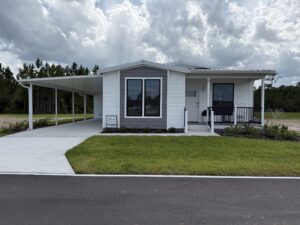 Silver Bay model manufactured home with a covered carport, front porch, landscaped yard, and wooded area behind it.