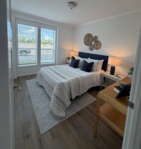 Manufactured home bedroom at Silver Bay with a queen-size bed, gray bedding, bedside lamps, sliding glass doors, and wood-look flooring.